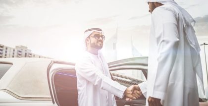 Two men in traditional attire shaking hands beside a car.