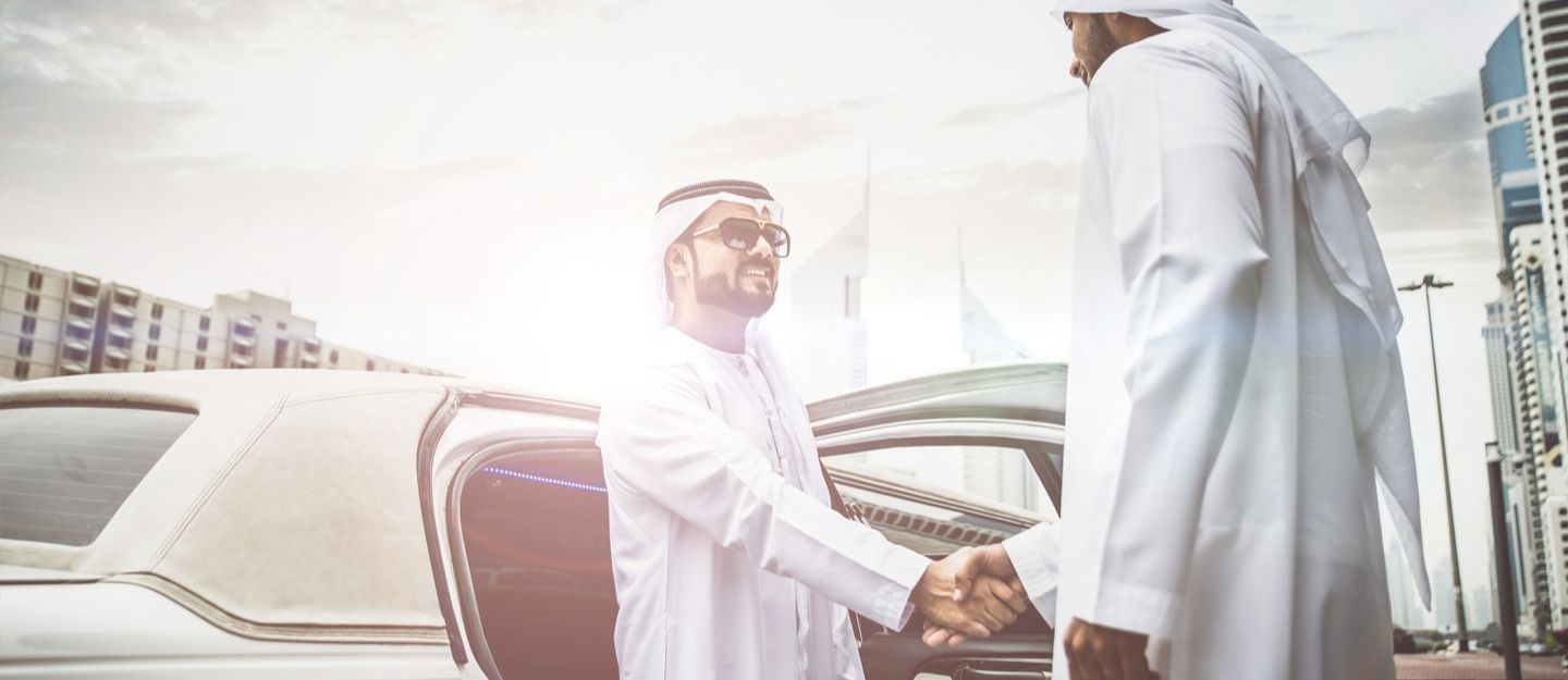 Two men in traditional attire shaking hands beside a car.
