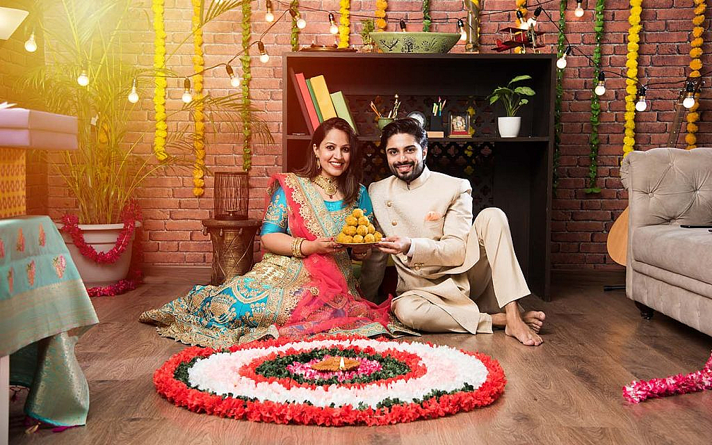 A couple sitting beside a rangoli and holding sweets on Diwali