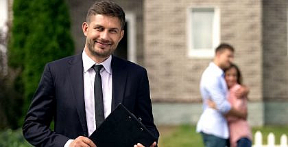 Licensed real estate broker in Dubai standing with documents and smiling, while a happy family sits in the background, celebrating their new home purchase.