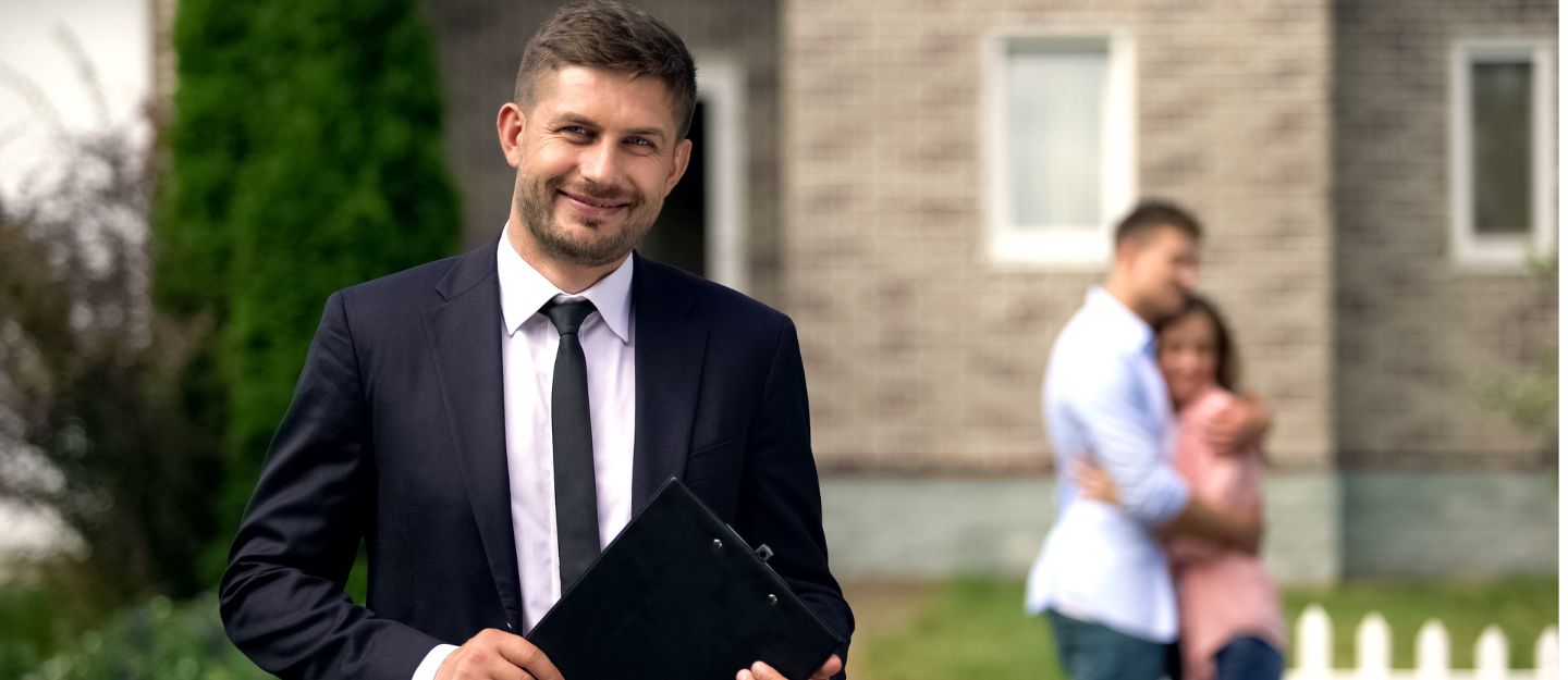 Licensed real estate broker in Dubai standing with documents and smiling, while a happy family sits in the background, celebrating their new home purchase.