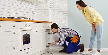 Plumber and woman inspecting a clogged drain under a modern white sink in an apartment kitchen.