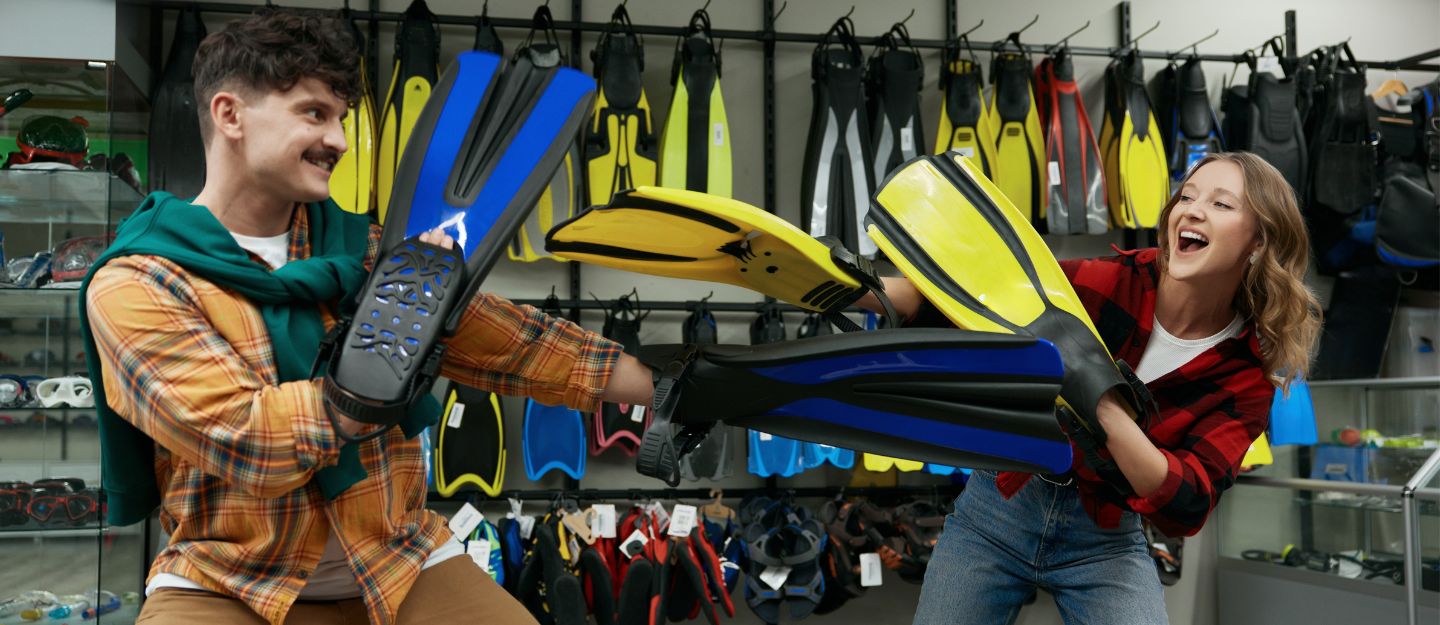 Man and woman playfully holding swimming fins in a swimming pool supply shop in Abu Dhabi, surrounded by displays of fins.