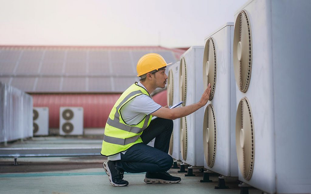 A man in a hard hat and safety vest is inspecting HVAC units on a rooftop.
