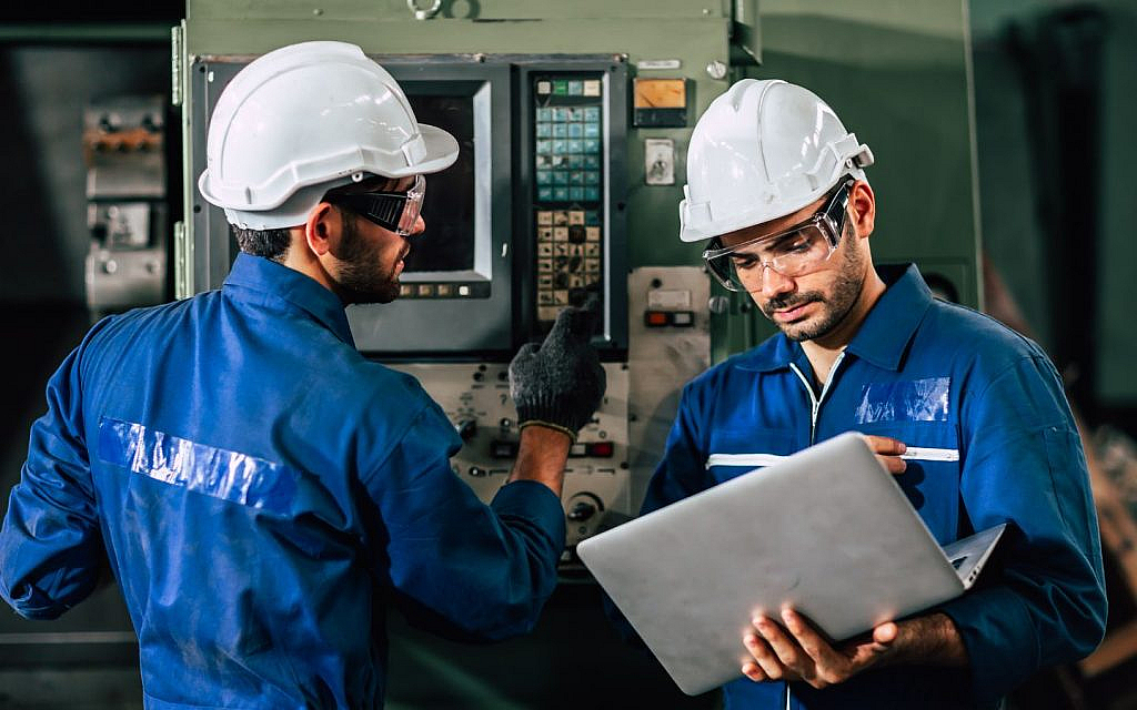 Two maintenance workers in hard hats and safety glasses are working with machinery and a laptop.

