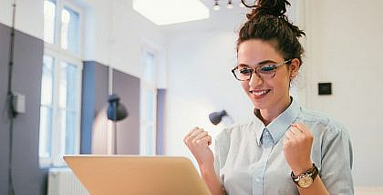 Woman smiling and making a celebratory gesture while looking at a laptop, representing the excitement of becoming a Real Estate Area Specialist