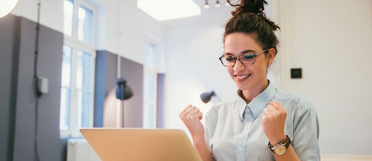 Woman smiling and making a celebratory gesture while looking at a laptop, representing the excitement of becoming a Real Estate Area Specialist