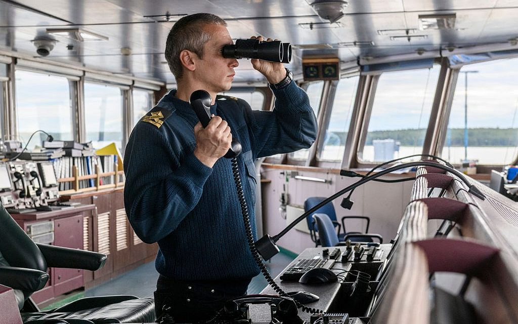 A ship captain is looking through binoculars and holding a telephone receiver on the bridge of a vessel.