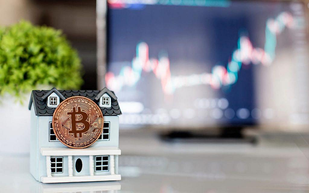 A miniature house with a Bitcoin coin leaning against it and a blurred computer screen in the background.