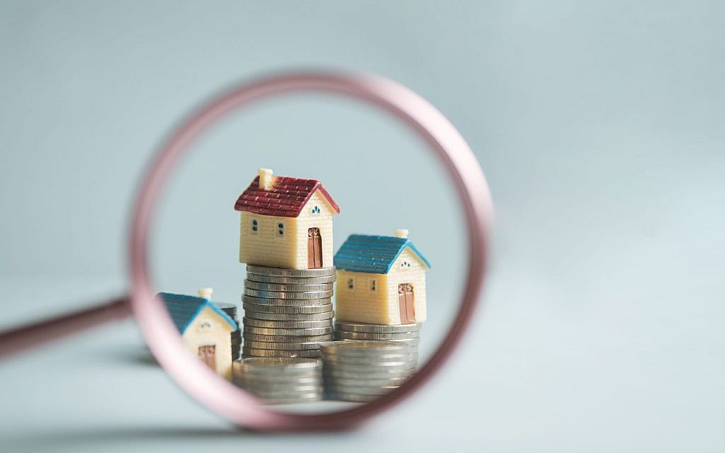 A magnifying glass focused on miniature houses stacked on coins, representing real estate valuation.