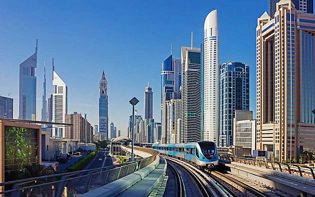 Dubai Metro train on an elevated track, passing by the city's modern skyscrapers under a clear blue sky.