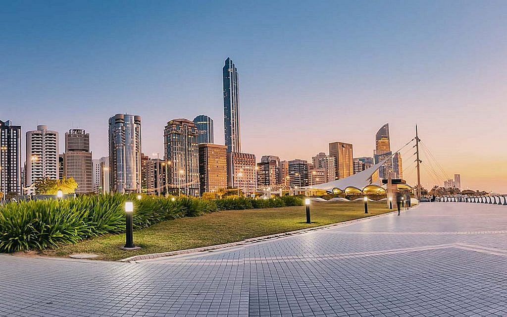 City skyline with modern buildings, waterfront promenade and illuminated green space at sunset.