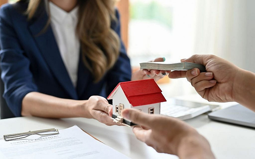 Hands exchanging a small house model and money over a desk with a contract.
