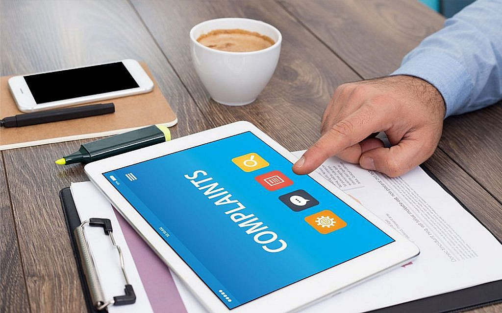 Hand on desk points to tablet displaying 'COMPLAINTS' and icons, next to phone and coffee cup.
