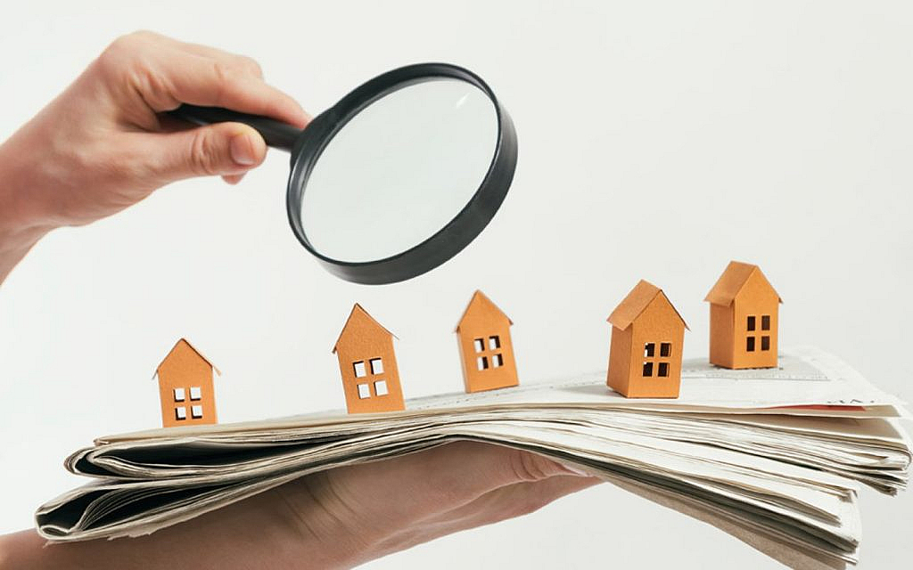 A hand holding a magnifying glass over miniature paper houses arranged on a stack of newspapers.
