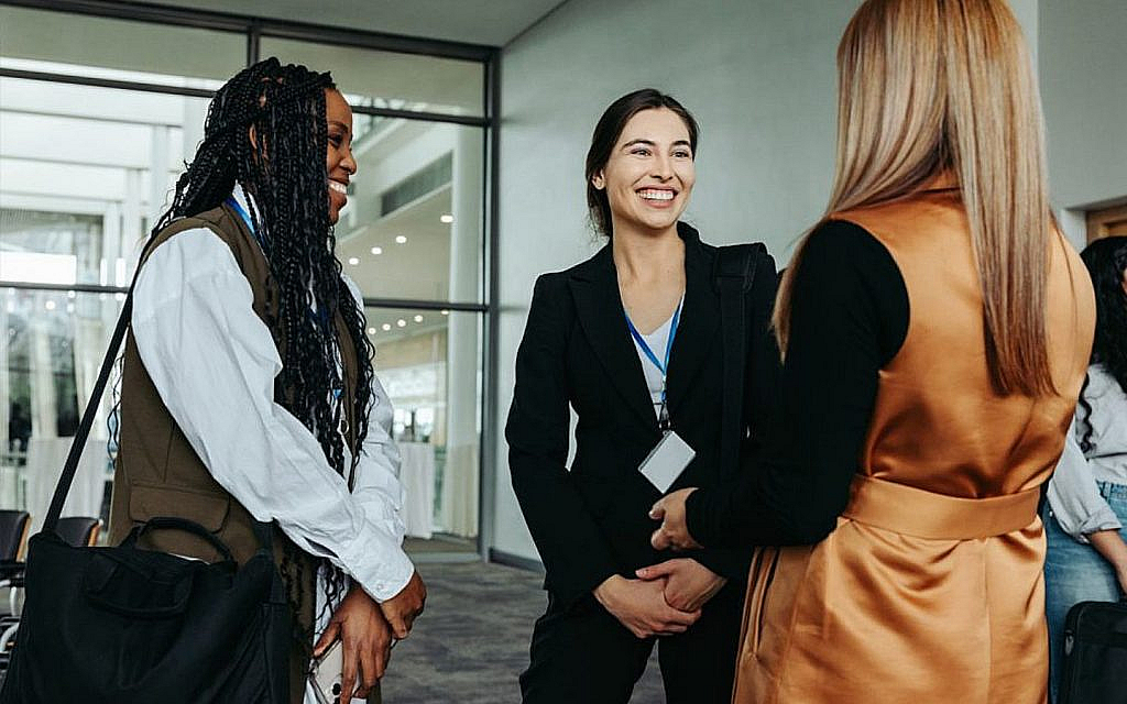 Three women smiling and conversing in a brightly lit modern interior, possibly at a conference or event.
