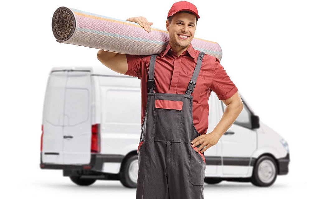 A male worker carrying a rolled-up carpet over his shoulder, with a cleaning van in the background.
