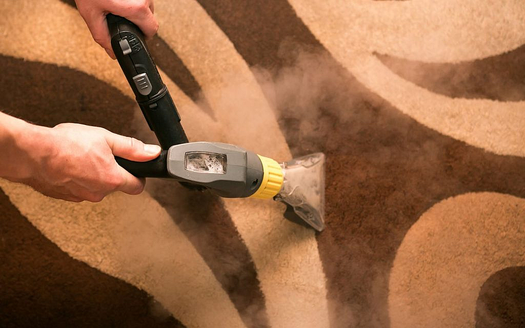Close-up of a person's hands using a steam vacuum cleaner to clean a patterned carpet.
