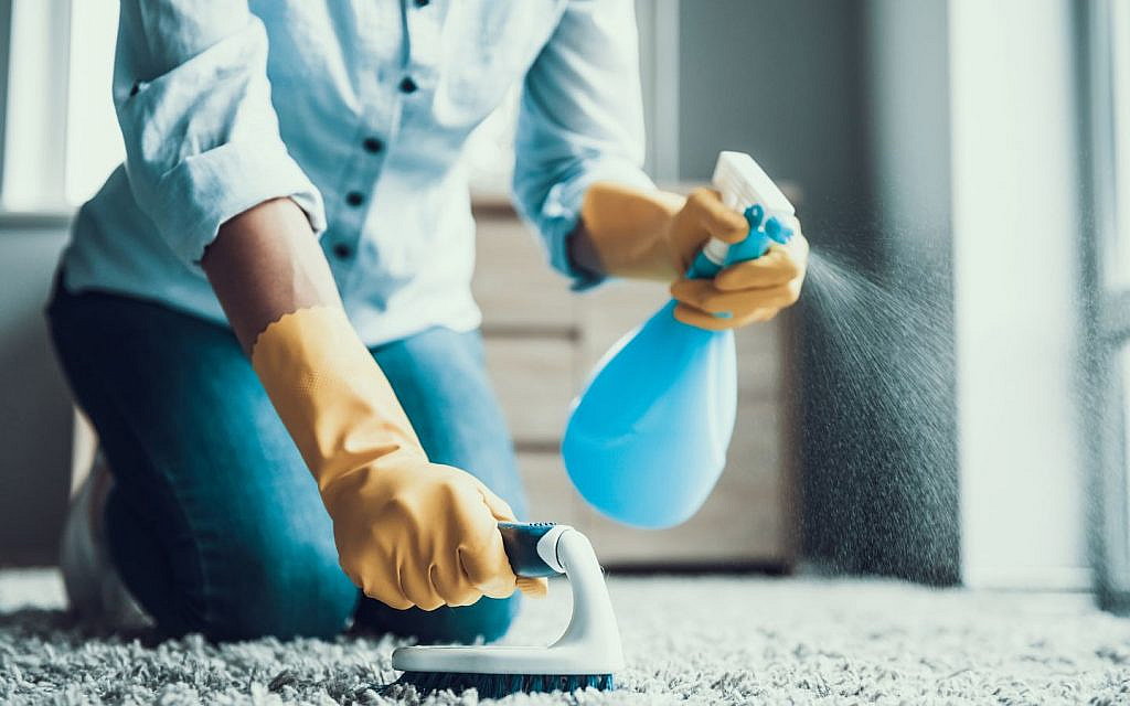 Person on their knees, spraying a carpet with a cleaning bottle and scrubbing it with a brush.
