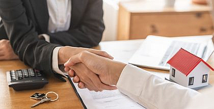 شخص يصافح شخص آخر على طاولة تحتوي مجسم منزل ومفتاح منزل وأوراق