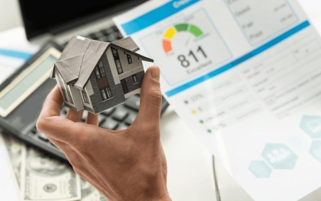 A person's hand holding a small model house in front of a credit report