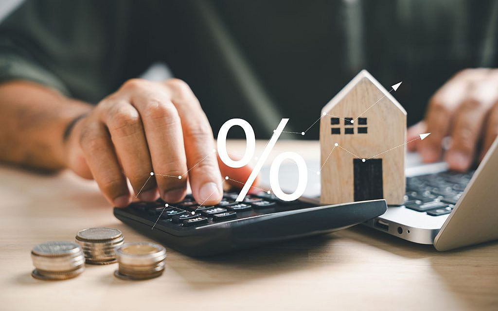 A person using a calculator and a laptop next to a small wooden house model and stacks of coins