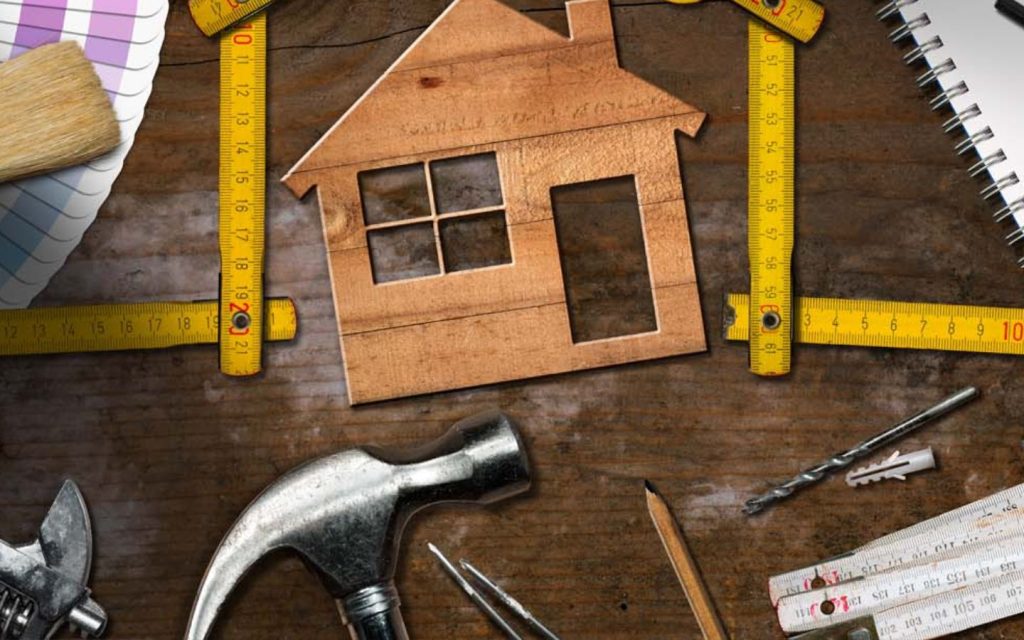 An overhead shot of various construction and design tools surrounding a small wooden house cutout.
